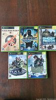 Seven Xbox game cases arranged on a wooden surface showing Red Faction II, James Bond 007 Agent Under Fire, Ghost Recon Island Thunder, Halo Combat Evolved, and Medal of Honor Frontline.