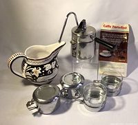 Photo showing the complete espresso set including the metal espresso maker, cups, container, ceramic decorative pitcher, and instruction leaflet