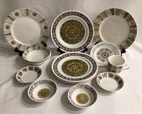 View of 14 Kathy Winkle ceramic pieces including plates, bowls, cups, and saucers arranged with two plates on stands and others laid out showing decorative rims and centers