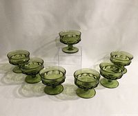 Seven green King's Crown Thumbprint sherbet glasses arranged in a semi-circle around one glass on a clear stand, showing color and quantity.