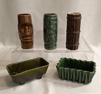 Front view showing three tiki cups elevated on a clear stand and two green planters below on surface