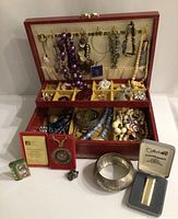 Photo of open vintage red jewellery box displaying assortment of necklaces, bracelets, rings, earrings, brooches, medallion, lighter, and pill container arranged inside and outside the box.