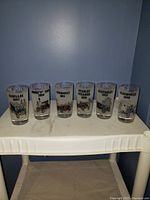 Six clear drinking glasses arranged in a row on a white surface against a gray wall, each glass printed with a different antique car and year.