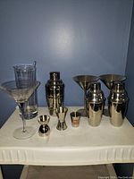 View of entire bar drinkware set showing multiple cocktail shakers, jiggers, shot glass, clear glass cups, and a metal martini glass-shaped cup arranged on a white surface against a plain blue wall.