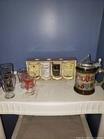 Overall view of the German beer stein, the set of five whiskey glasses, and individual shot glasses on a shelf.