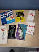 Image showing six bartending books matching various cocktail and bartending topics plus two white Budweiser branded boxes likely containing cocktail shakers or related bar tools.