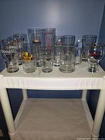Multiple beer glasses of various brands arranged on a white plastic shelf against blue wall