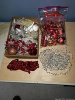 Photo showing box with assorted Christmas ornaments including red bows, silver stars, glittery baubles, bead garlands in red and silver, and white frosted clusters.