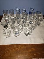 Lot of assorted clear glassware including shot glasses, mugs, and a square decanter on a white cloth.