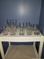 14 assorted clear drinking glasses on a white surface against a blue wall, showing a mix of tall and short styles including cut crystal patterns