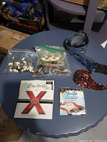 Photo showing two packs of guitar strings, two bags of cream colored tuning pegs, blue guitar strap and tortoiseshell pick on blue table.