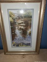 Full view of framed landscape print depicting a water scene with reflection under a bridge, inside a gold-tone frame with white and beige matting.