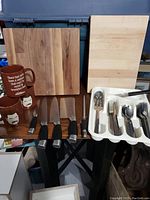 Two wooden cutting boards, knives, three brown mugs, and a plastic tray with assorted flatware and utensils displayed on a table