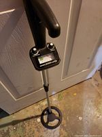 Metal detector close-up showing control panel with analog meter, power and sensitivity knobs, and armrest handle.