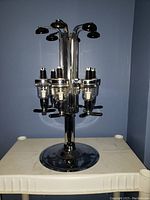 Front view of chrome rotating bottle dispenser with six pour spouts and pedestal base on white table against blue wall.