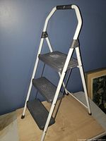 Full side view of the Gorilla 3-step metal ladder with white frame and black steps on a wood floor against a blue wall