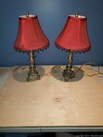 Pair of ornate brass table lamps with red fabric shades featuring bead trim on a light wooden surface against a blue wall.