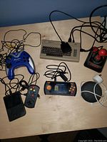 Overview photo showing various handheld game consoles, controllers, vintage keyboard, cables, and accessories on a wooden surface.