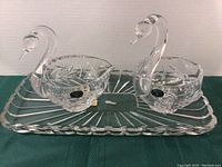 Two crystal swan serving bowls and a rectangular crystal drip tray arranged on a table. Both swan bowls have clear pinwheel cut patterns and original Bohemia stickers visible on the bowls. The drip tray has a scalloped edge and cut linear design.