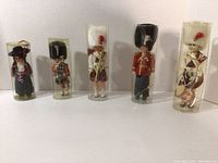 Full group of five vintage Royal Guard dolls and one girl doll in original cylindrical plastic containers; three 9-inch dolls missing lids, one 7-inch doll and one girl doll with lids.