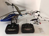 Side view of large blue Protocol helicopter with carbon fiber style canopy and structural frame, smaller white and blue XL Litehawk helicopter, and two black remote controls with labeled buttons and sticks.