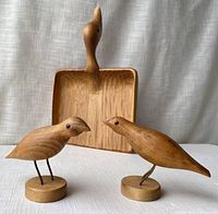 Front view of wood carved bird dust pan behind two wood bird figurines with glass eyes and metal legs, showing the natural wood color and grain