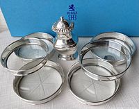 Six round sterling silver coasters with clear glass bottoms and one sterling silver shaker with perforated lid displayed with Birks blue box in background.