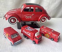 Four red Coca-Cola branded die-cast model vehicles on white textured fabric background including a VW Beetle and three smaller vehicles.