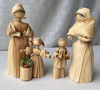 Front view of four cornhusk dolls: two mother dolls and two child dolls, including the girl holding a basket with produce.