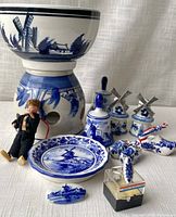 Porcelain items arranged on white fabric background showing bowl on planter, doll, dish, windmill shakers, bells, brooch, ring, and Dutch shoes.
