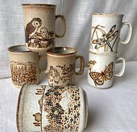 Six Dunoon ceramic mugs arranged in stack and lying on side, showing detailed nature and animal designs including owls, butterfly, dragonfly, floral scenes.