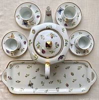Top view showing the teapot, four petite cups on saucers arranged around it, and rectangular serving tray below with butterfly and floral design and gold trim.