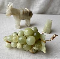 Photo showing the onyx donkey figurine, grape bunch with metal stem, and small carved figure on a white cloth background.