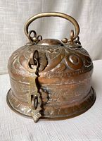 Copper coated metal circular container with hinged lid and handle, engraved decorations visible, latch closed, shown on fabric background.