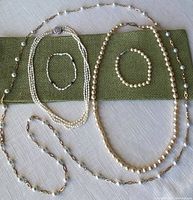 Four necklaces and three bracelets arranged on green fabric and white tablecloth, showing size and style variation including costume and freshwater pearls.