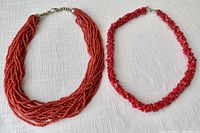 Two red coral necklaces laid flat on a white textured fabric background, showing the overall design and length.