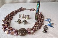 Lot arranged to show whole set including tripple strand shell necklace with clasp, brass and shell bangle, three pairs of earrings, and dark pearl pendant on white woven fabric.
