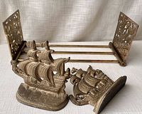 Two solid cast metal ship-shaped bookends and a pair of folding metal bookends shown on a fabric surface. The ship-shaped bookends have detailed sails and hulls with an aged patina. The folding bookends feature decorative perforated metal ends and three metal rods connecting them.
