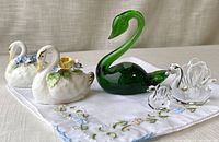 Green handblown glass swan, two clear glass swans, two Adderly England porcelain swans with small flower decorations, displayed on embroidered linen cloth.