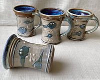 Four beige ceramic mugs decorated with blue and green tadpole-like figures, showing front and side views with textured surface and glossy dark blue interior.