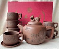 Front side view of the full set showing teapot with six stacked cups and saucers in front of red box with Chinese gold lettering.