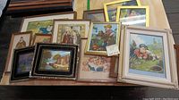 Photo of a group of 12 framed prints and paintings stacked, showcasing varied subjects including children, babies, religious scenes, still life theorem paintings and scenic images.