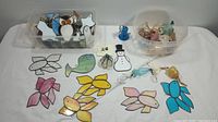 Photo of various stained glass ornaments including owls, fish, balloons, ghosts, flowers, and butterflies laid out on a white surface in two clear plastic containers and loose on the table.