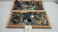 Overview of mixed costume jewelry in two wooden trays, showing a large assortment of bracelets, brooches, necklaces, and earrings.