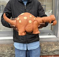 Front view of the brown dinosaur rubber plush with cream spots, held in adult hands illustrating size and texture.