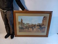 Full view of framed lithograph held by person for scale, showing artwork and frame condition.