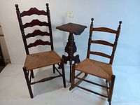 Front view showing two ladder back wooden chairs with woven rush seats, one rocking chair and one stationary, alongside a wooden plant stand with a square top and tripod carved base