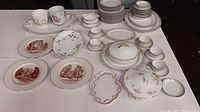 Photo showing an assortment of china and transferware plates, serving pieces, and teaware arranged on a white surface.
