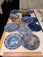 Top-down photo showing 15 vintage ironstone transferware plates in blue and brown on a white surface with a ruler and apple for scale.