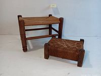 Two wooden footstools with woven rush seats, placed on white wood floor against white background. Larger footstool has diagonal woven pattern with round legs, smaller footstool has checkerboard woven pattern with block legs.